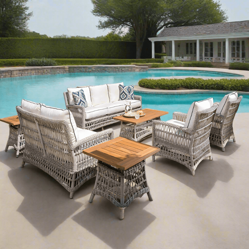 A seven-piece outdoor conversation set with white woven vinyl seating and brown teak wood table tops, placed on a patio near a pool.