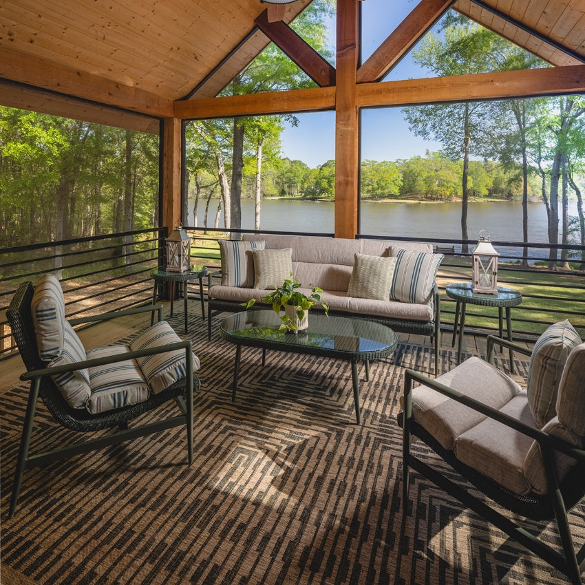A screened-in porch living room with a glass-top sofa, two cushioned chairs, an oval cocktail table, and a patterned rug, overlooking a lake and forest view.