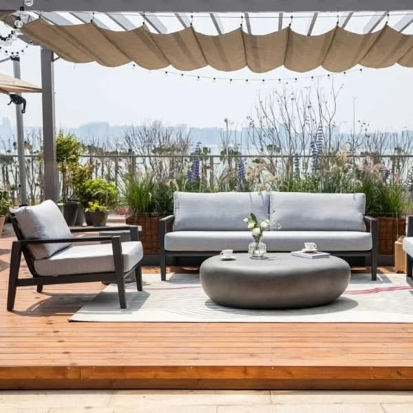 Outdoor patio furniture set on a wooden deck under a beige canopy, featuring a gray three-seat sofa, matching lounge chair, round coffee table, and a light rug, with potted plants and a distant garden view.