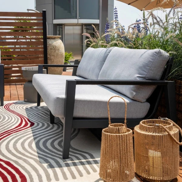 A modern outdoor patio sofa with a black aluminum frame and light gray cushions, styled on a patterned rug with woven lanterns and plants in the background.