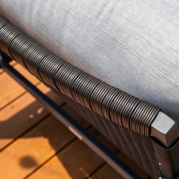 Close-up of a patio swivel glider with a light gray textured cushion, dark gray woven armrest, and a wooden deck floor in the background.