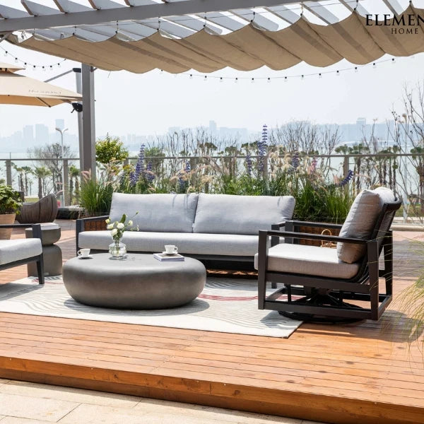 Outdoor patio furniture set including a light gray sofa, matching swivel glider chair, round coffee table, and a patterned area rug on a wooden deck under a beige canopy. The brand name 'ELEMENTI HOME' is visible in the top right corner.