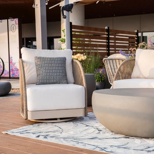 A modern outdoor patio chair with white cushions and a gray patterned accent pillow, set on a wooden deck next to a round coffee table and wicker seating.