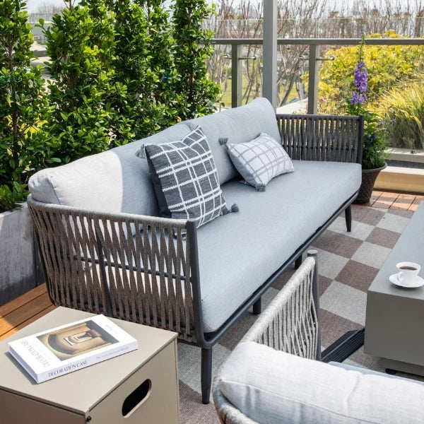Outdoor patio sofa with woven gray frame and light gray cushions, styled with two patterned throw pillows on a balcony with greenery and a coffee table nearby.