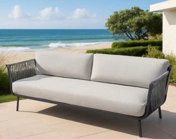 A modern outdoor sofa with light beige cushions and a dark gray woven frame, positioned on a tiled patio with a beach and ocean background.