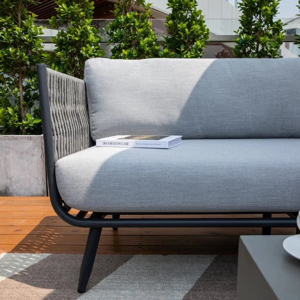 A modern outdoor sofa with light gray textured cushions and a black metal frame, shown on a wooden deck with green plants in the background. A book rests on the seat cushion.