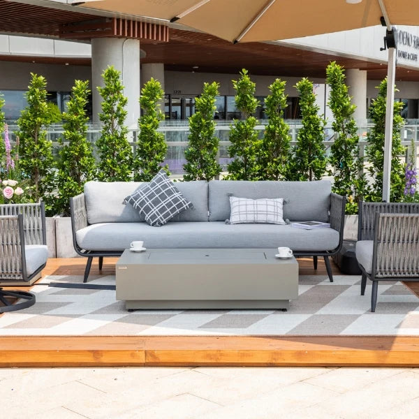 A modern outdoor patio furniture set featuring a light gray three-seater sofa with two patterned throw pillows, a matching rectangular coffee table, and two armchairs on a checkered rug. The set is styled on a wooden deck with a patio umbrella, green shrubs, and a building in the background.