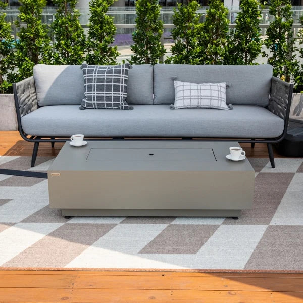 A modern outdoor patio scene featuring a gray sectional sofa with two patterned throw pillows and a matching rectangular coffee table in the foreground, set on a geometric rug with a wooden deck and green shrubs behind.