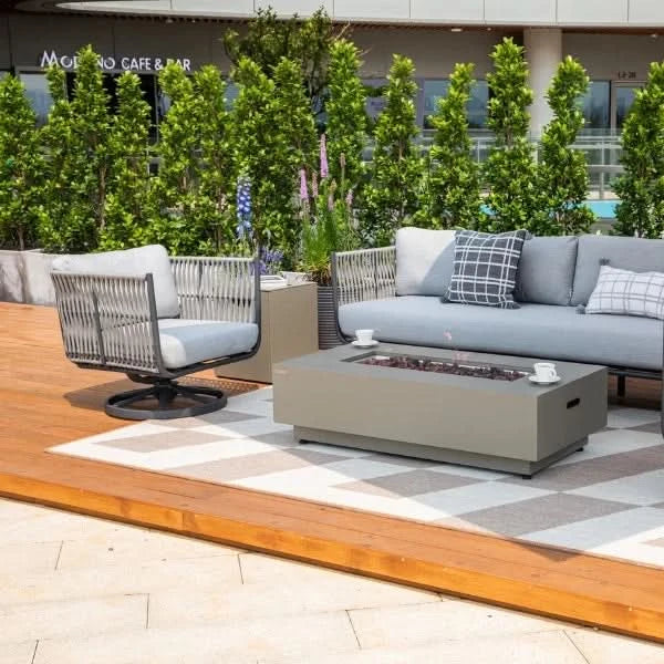 Outdoor patio furniture set including a light gray sectional sofa, matching armchair, rectangular fire pit table, and a patterned area rug on a wooden deck with greenery in the background.