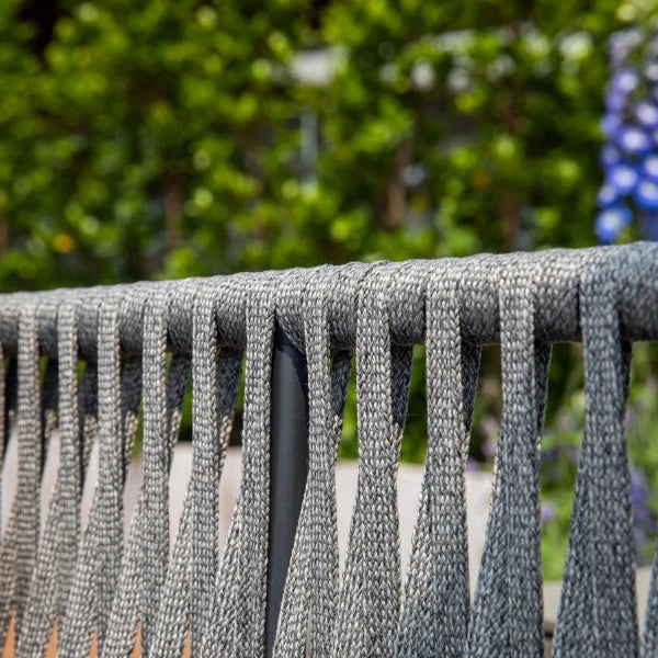 Close-up of a gray woven outdoor chair sling with vertical straps hanging from a metal frame, set against a blurred green garden background.