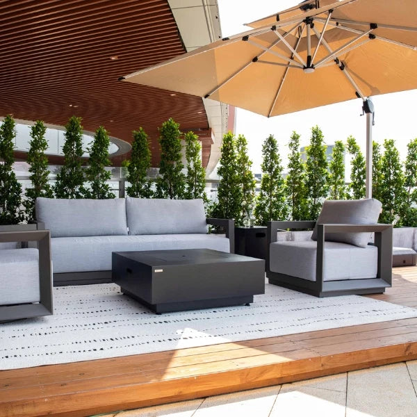 Modern outdoor patio furniture set including a light gray sofa, matching lounge chair, dark gray coffee table, and a large beige patio umbrella on a wooden deck.