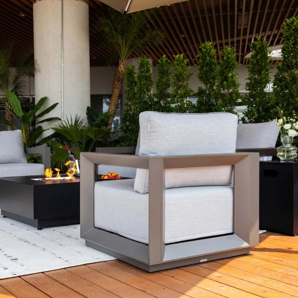 A modern outdoor patio armchair with a light gray textured fabric cushion and a square metallic frame, shown on a wooden deck next to a black fire pit table.
