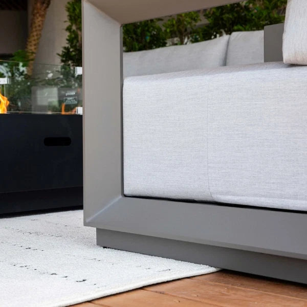 Close-up of a modern outdoor lounge chair with light gray cushions and a dark gray frame, shown on a patio with a white rug, wood floor, and a black side table nearby.
