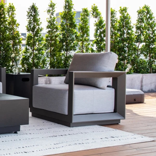 A modern outdoor lounge chair with a dark gray frame and light gray cushion, shown on a wooden deck with a white rug, potted trees, and a patio umbrella in the background.