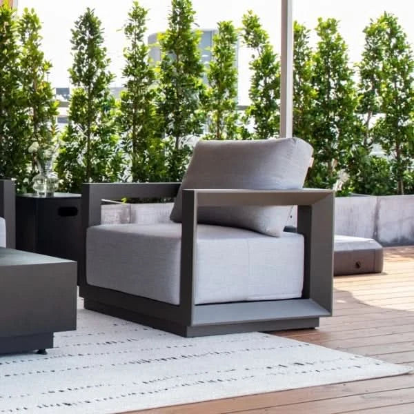 A modern outdoor lounge chair with a dark gray frame and light gray cushion, shown on a wooden deck with a white rug, potted trees, and a patio umbrella in the background.