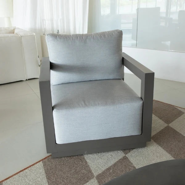 A modern outdoor lounge chair with a light gray textured fabric cushion and a dark gray frame, positioned on a checkered rug in a bright, modern living space.