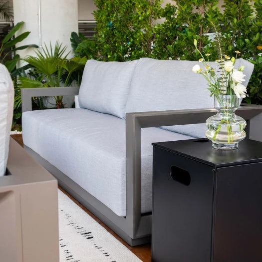A modern outdoor sofa with light gray cushions and a dark gray frame, shown in a garden setting next to a black side table with a glass vase of white flowers.