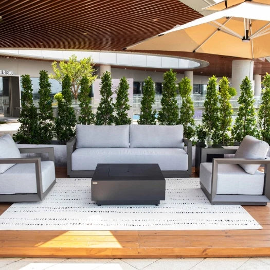 Modern outdoor patio furniture set including a three-seat sofa, two armchairs, and a rectangular coffee table on a white patterned rug, with tall green plants in the background.