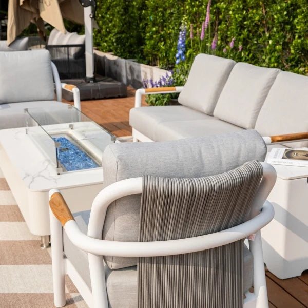 Outdoor patio furniture set with a white-framed seating area featuring light gray cushions, a matching fire pit table, and a striped fabric backrest, set on a wooden deck with greenery in the background.
