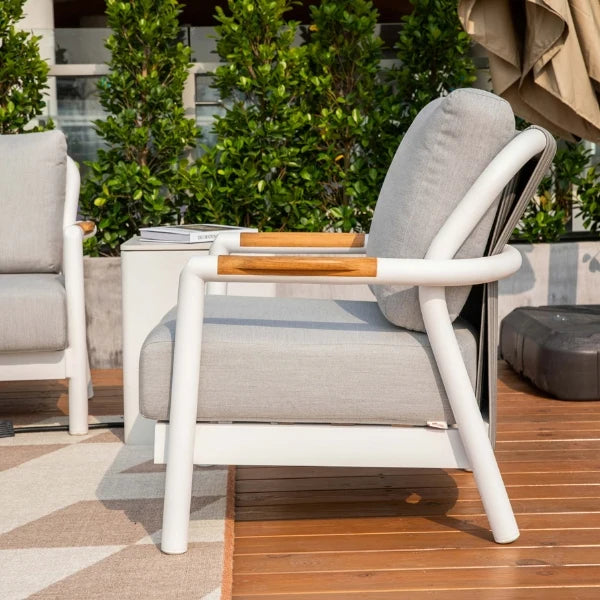 A modern outdoor armchair with a white frame, light gray cushioned seat and back, and light wood armrest accents, shown on a wooden deck with greenery in the background.