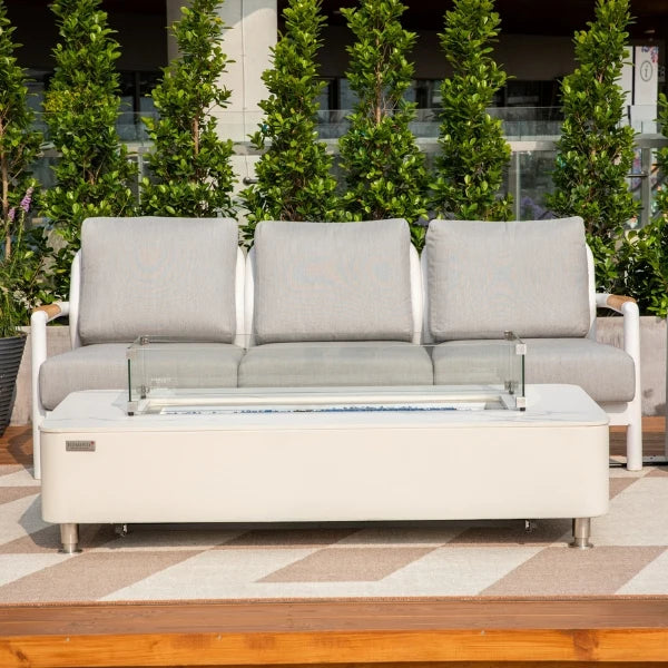 Outdoor patio furniture set featuring a three-seat sofa with light gray cushions and a matching rectangular fire pit coffee table, set on a patterned rug with green shrubs in the background.