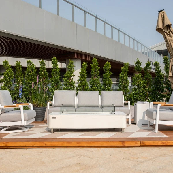 A modern outdoor patio furniture set featuring a three-seat sofa, matching armchair, side table, and a low rectangular coffee table, all in white frames with light gray cushions, arranged on a patterned rug in front of a contemporary building.