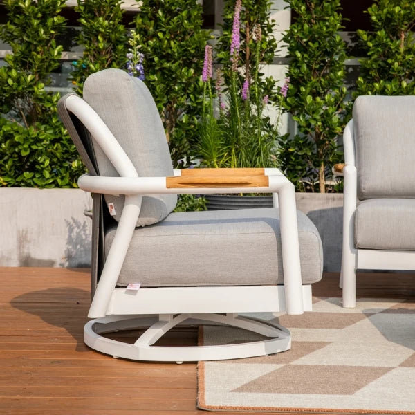 A modern outdoor swivel lounge chair with light gray cushions, a white frame, and a light wood armrest, shown on a wooden deck with green plants in the background.