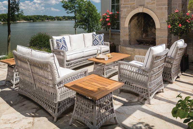 A seven-piece outdoor conversation set with white woven vinyl seating and brown teak wood table tops, placed on a patio near a pool.