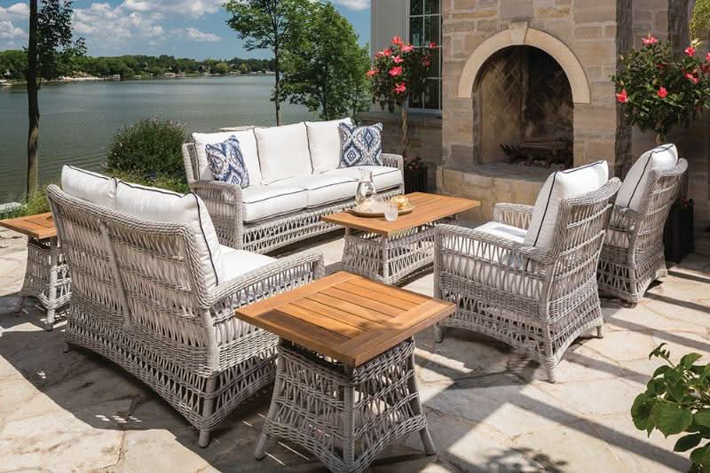 A seven-piece outdoor conversation set with white woven vinyl seating and brown teak wood table tops, placed on a patio near a pool.