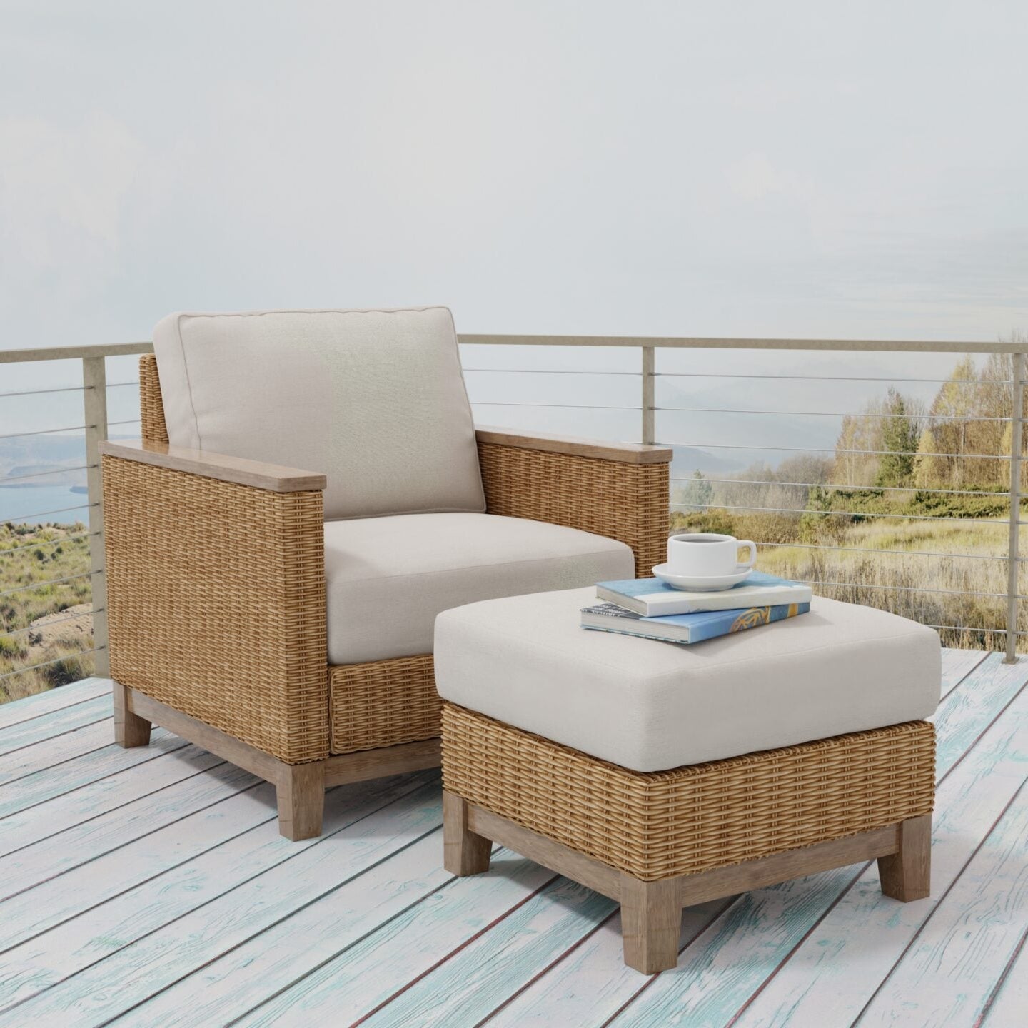 A wicker outdoor armchair with a light beige cushion sits on a light wood deck beside a matching ottoman with a white cushion. A small stack of books and a cup are placed on the ottoman, with a railing and a scenic landscape in the background.