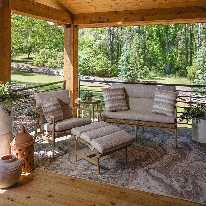 A 3-piece outdoor patio furniture set including a loveseat, armchair, and ottoman, styled on a patterned area rug on a wooden covered porch with a lush green forest background.