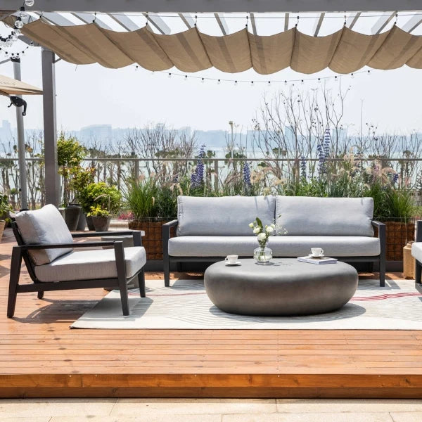 Outdoor patio furniture set on a wooden deck under a beige canopy, featuring a gray three-seat sofa, matching lounge chair, round coffee table, and a light rug, with potted plants and a distant garden view.