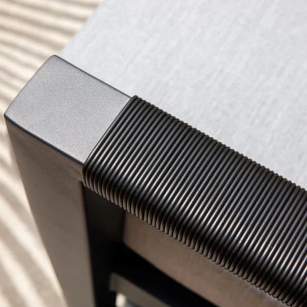 Close-up detail of a dark gray aluminum chair corner with a ribbed armrest and a light gray upholstered seat, shown in bright natural light.