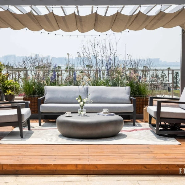 Outdoor patio furniture set including a gray three-seat sofa, matching armchair, and a round dark coffee table on a light rug, arranged on a wooden deck under a beige canopy with a scenic background.