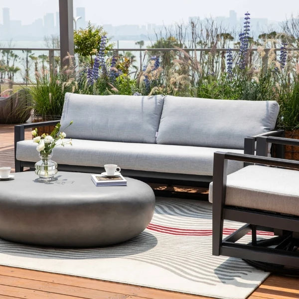 Outdoor patio furniture set featuring a light gray fabric sofa, a matching armchair with a cushion, a round dark gray coffee table, and a striped area rug on a wooden deck with a city skyline and garden in the background.