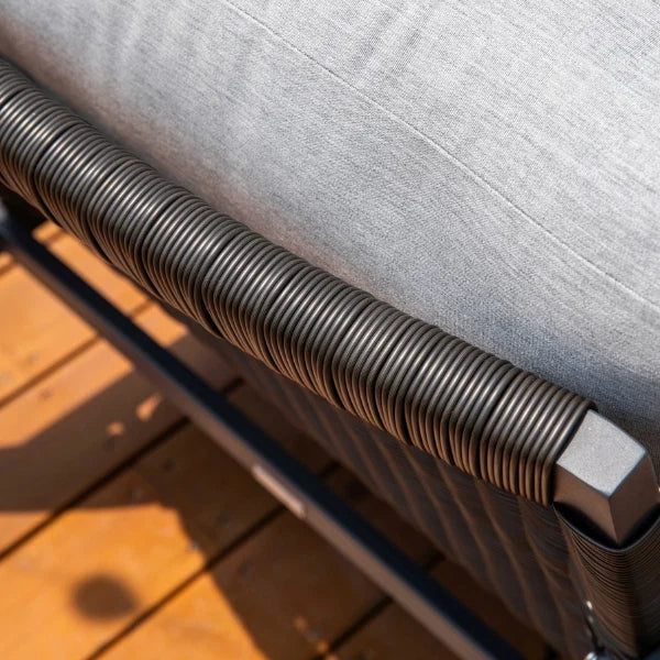 Close-up of a patio swivel glider with a light gray textured cushion, dark gray woven armrest, and a wooden deck floor in the background.