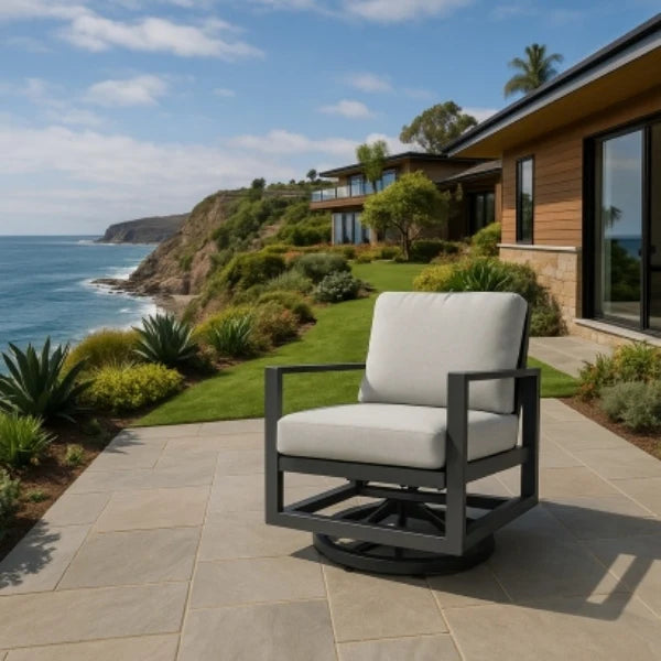 A modern outdoor swivel chair with a dark gray metal frame and light beige cushions, positioned on a stone patio overlooking a scenic coastal landscape with ocean, grass, and a house.