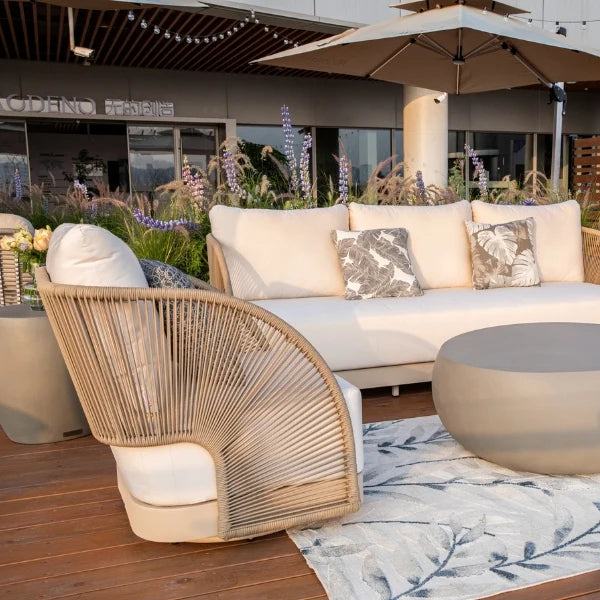 Outdoor patio furniture set featuring a curved woven rattan armchair with white cushions, a matching white sectional sofa with patterned accent pillows, a round concrete coffee table, and a leaf-patterned rug on a wooden deck.