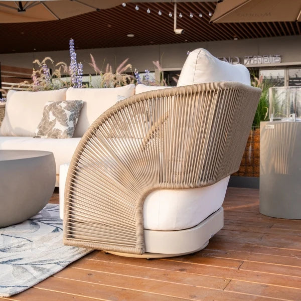 A modern outdoor swivel lounge chair with a curved back made of woven natural-fiber rope and thick white cushions, shown on a wooden patio next to a white sofa.