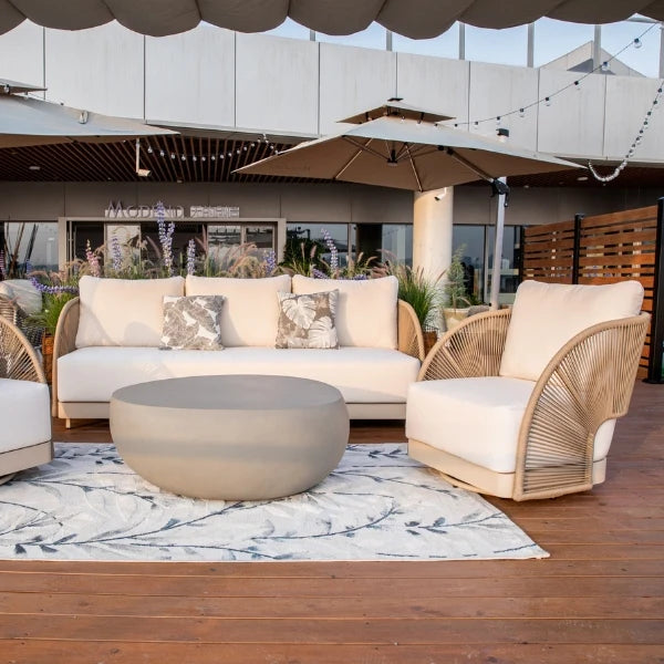 Outdoor patio furniture set including a cream sofa, two wicker armchairs with cream cushions, a round concrete coffee table, and a patterned area rug on a wooden deck.