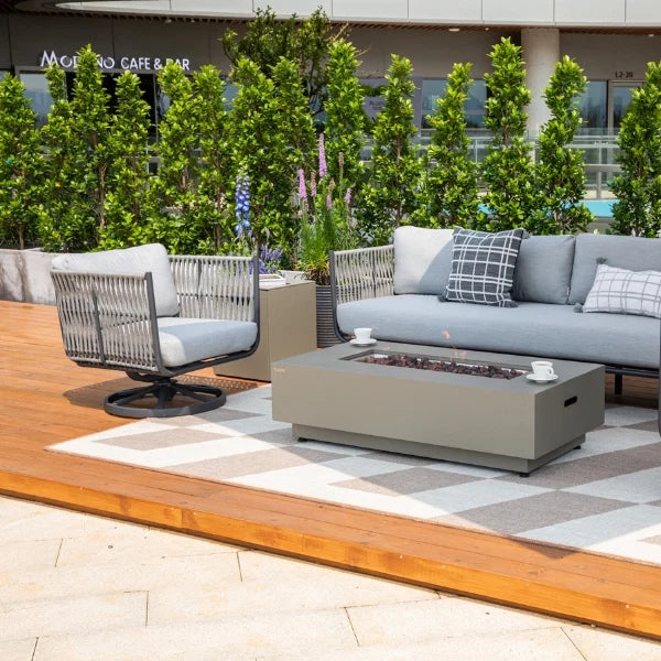 Outdoor patio furniture set including a light gray sectional sofa, matching armchair, rectangular fire pit table, and a patterned area rug on a wooden deck with greenery in the background.