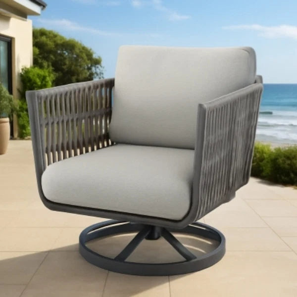 Outdoor swivel patio chair with a gray woven frame and light beige-gray cushions, shown on a tiled patio with a beach and ocean in the background.