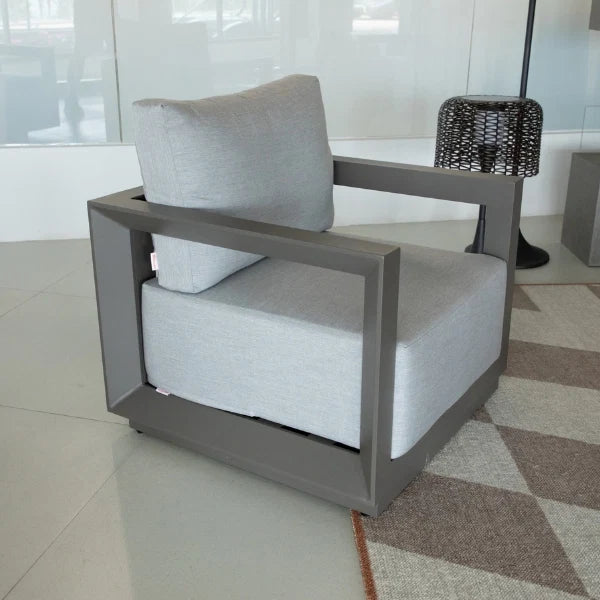A modern outdoor lounge chair with a light gray fabric cushion and a dark gray geometric frame, shown on a patterned rug in a bright, modern interior.