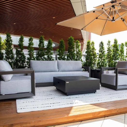 Modern outdoor patio furniture set including a light gray three-seater sofa, two matching armchairs, and a dark gray rectangular coffee table on a white patterned rug. The patio features a wooden slatted ceiling, a large beige patio umbrella, and a row of tall green plants in the background.