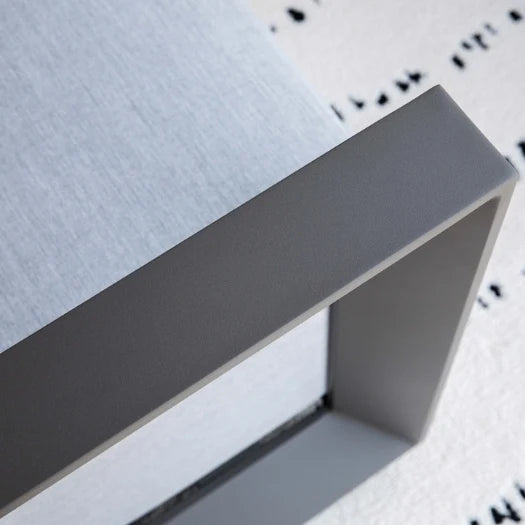 Close-up detail of a corner of the armrest of a modern outdoor sofa frame in matte gray, showing the corner joint and textured light gray fabric seat on a white patterned rug.