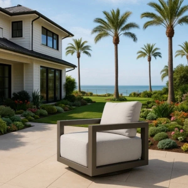 Modern outdoor swivel chair with white cushions in a garden setting with palm trees and a house.
