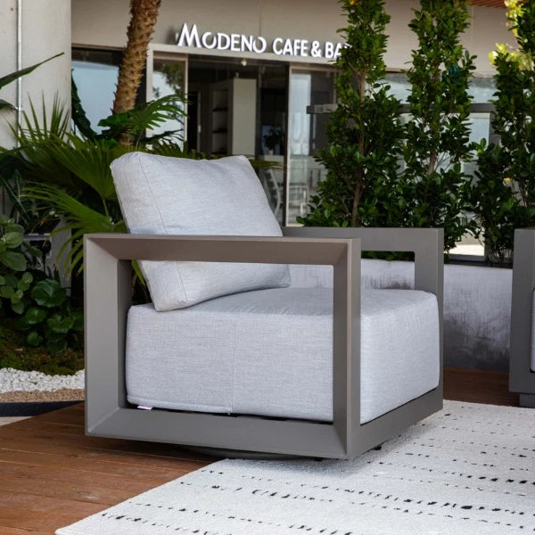 A modern outdoor swivel chair with a light gray textured fabric cushion and a square gray frame, placed on a white patterned rug in front of a cafe patio with green plants.