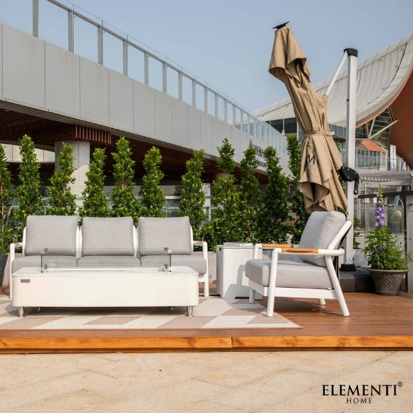 Modern outdoor patio furniture set by Elementi Home, including a white three-seat sofa with light gray cushions, a matching armchair, a low white coffee table, and a closed beige patio umbrella on a wooden deck.