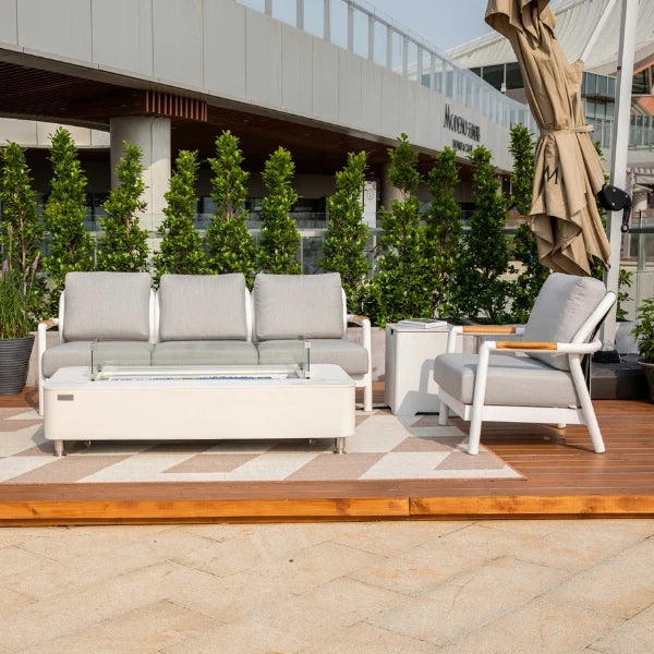 Modern outdoor patio furniture set including a three-seat sofa, armchair, and rectangular coffee table on a wooden deck with a patterned rug, greenery, and a building facade in the background.