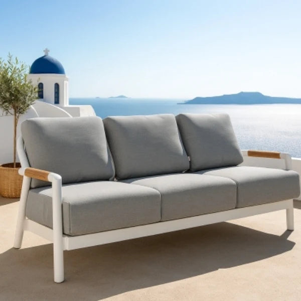 A three-seat outdoor sofa with light gray cushions and a white frame, styled on a patio with a blue-domed building and sea in the background.